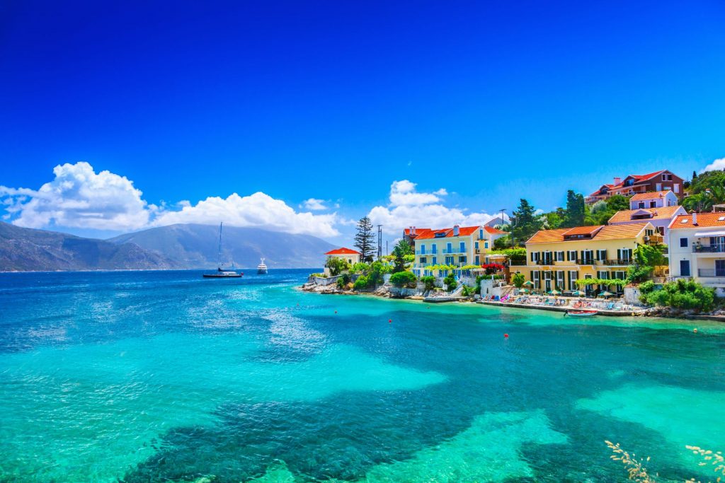 Turquoise water at Fiskardo, with colorful houses on the shore and blue sky. Sailboat on the horizon.
