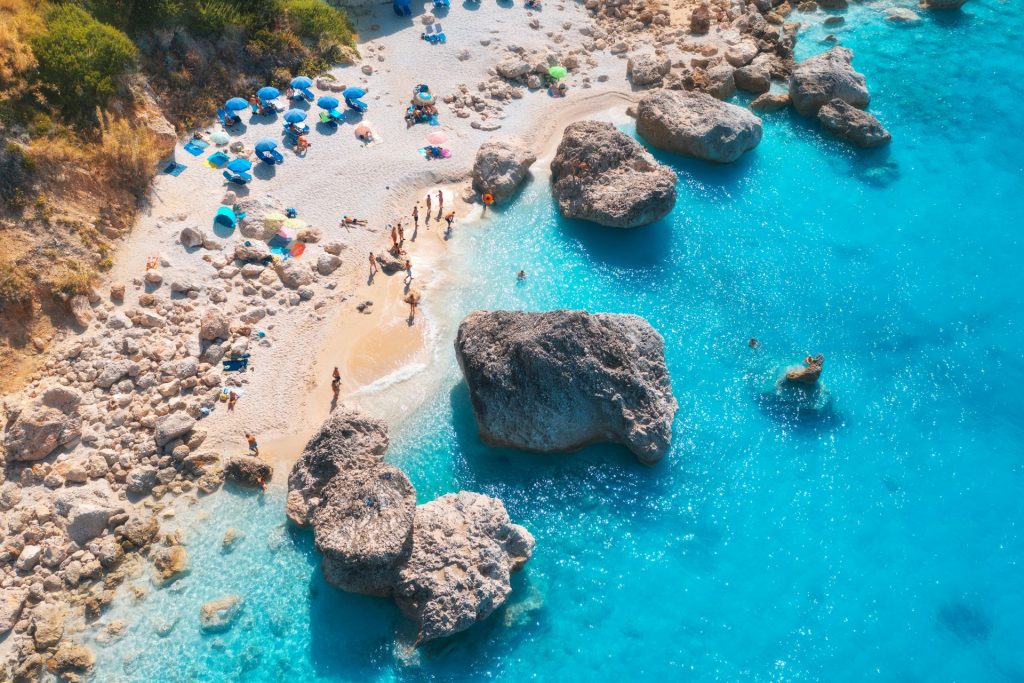Aerial of beach at Lefkada in Greece, with white sand and big rocky boulders. Blue beach umbrellas on the beach and people.