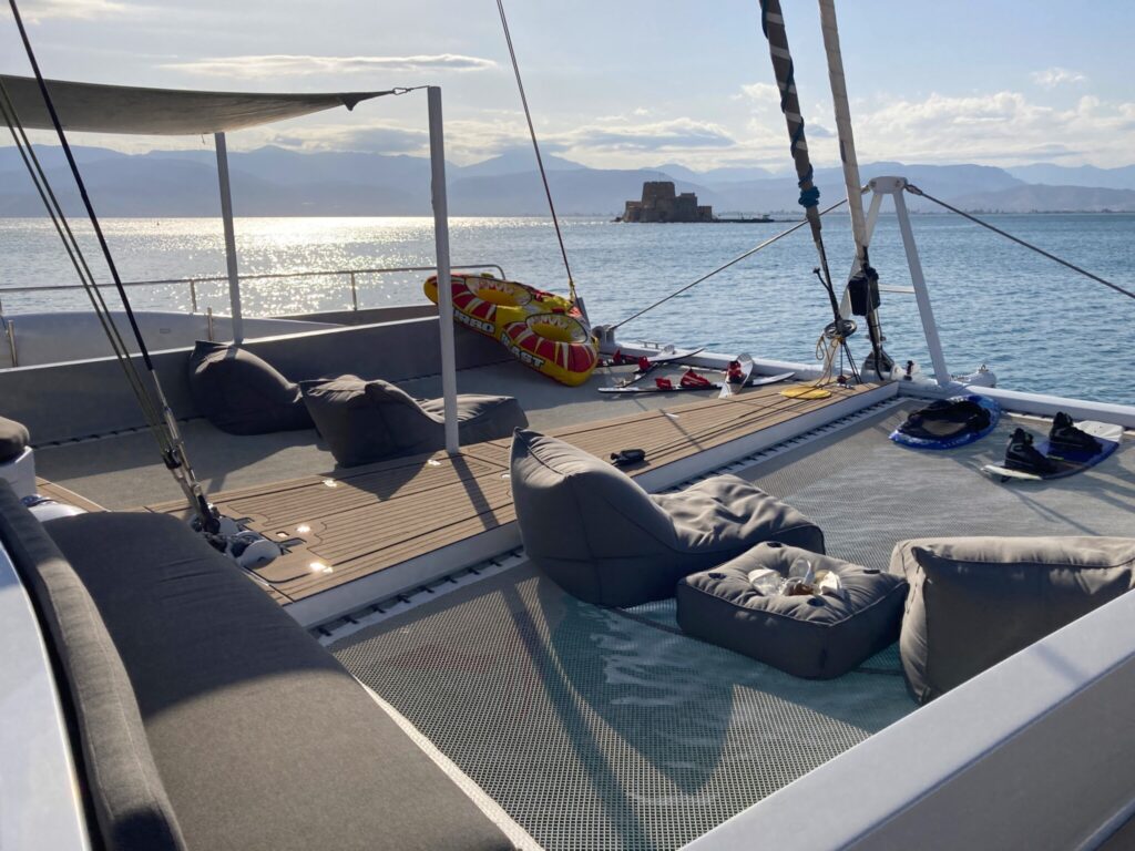 The teak foredeck of a 75-foot charter catamaran with sunbeds and water toys, Nafplio behind