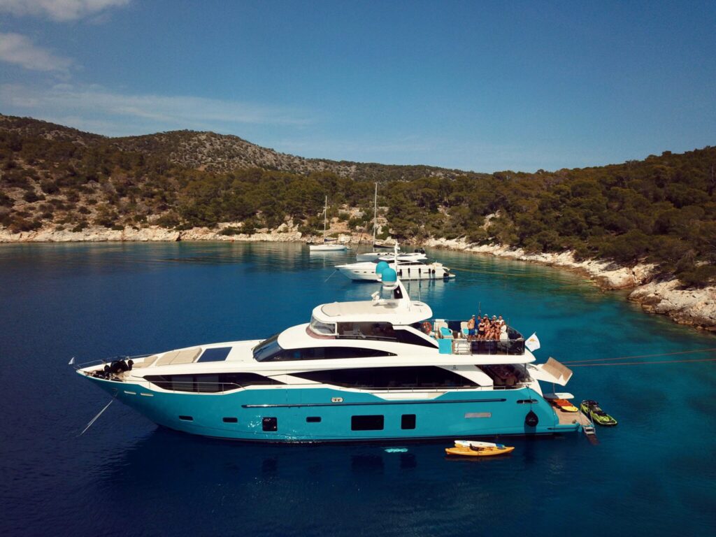 Motor yacht ANKA tied stern-to the rocks in Mouza Bay, Agistri, with guests waving from the aft deck, May 2025