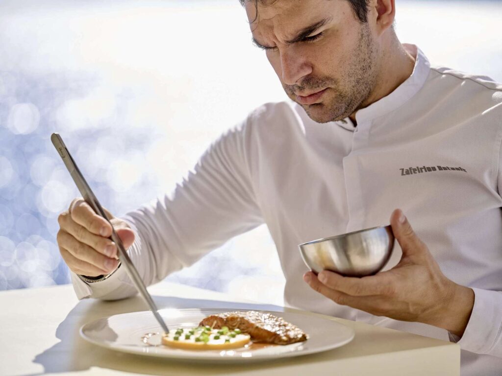 The chef of superyacht LA PELLEGRINA plating a dish with tweezers, sea light behind him