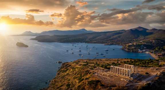Panoramic view of the Temple of Poseidon at Cape Sounion at the edge of Attica, Greece, with moored sailboats in the bay during sunset time