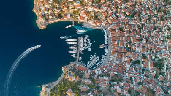 Aerial view of Hydra with many luxury yachts docked in the Mariana, which is situated in a snug little bay surrounded by red-roofed houses.