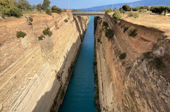 Daytime view of the Corinth Canal in Greece, seen from above. The canal is narrow with turquoise blue waters, flanked by tall, steep, sandy-colored cliffs. Sparse vegetation dots the cliff sides. A bridge spans across the canal in the distance, under a clear blue sky.