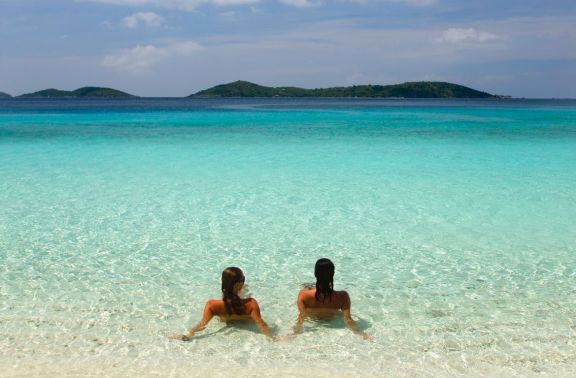 Two topless women sitting in the clear blue water on beach in Greece with their backs turned