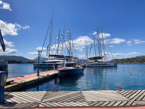 A vibrant scene at the yacht show in Poros, Greece, featuring a collection of sleek, modern yachts moored in the harbor. The backdrop showcases the charming coastal town with its traditional architecture, nestled against lush green hills under a clear blue sky. Crowds of visitors can be seen exploring the waterfront, adding a lively atmosphere to this picturesque maritime event.