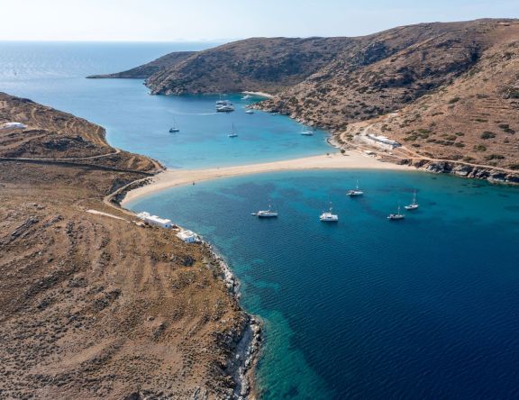 View of Kolona Beach on Kythnos, featuring a narrow isthmus of golden sand flanked by azure waters with yachts moored on both sides under a clear blue sky.