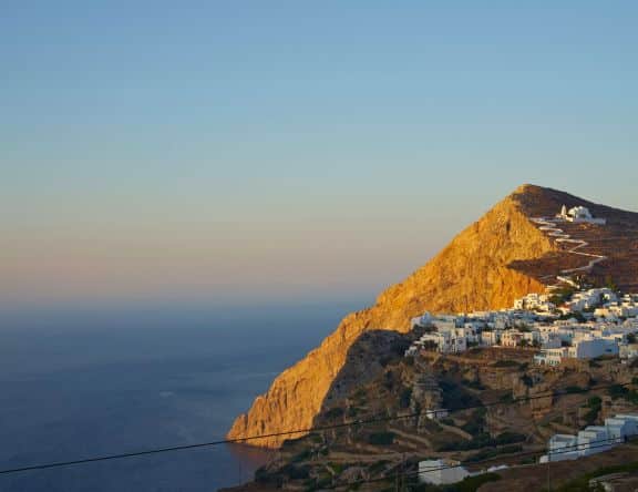 A panoramic view of Folegandros, Greece during sunset. The image captures the rugged, mountainous landscape bathed in golden light, highlighting the traditional white buildings scattered along the sloping terrain. The calm sea extends into the horizon under a clear sky, and a distinct church with a prominent white dome sits atop the hill, overseeing the scenic village below.