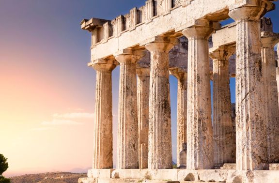 This image shows the ancient Greek temple of Aphaia on the island of Aegina, basking in the warm glow of a sunset. The Doric columns of the temple stand prominently against a pastel sky, overlooking a rugged landscape that stretches into the distance. This image shows the ancient Greek temple of Aphaia on the island of Aegina, basking in the warm glow of a sunset. The Doric columns of the temple stand prominently against a pastel sky, overlooking a rugged landscape that stretches into the distance.