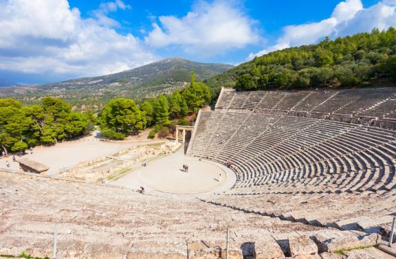 The ancient Epidaurus Theatre in Greece, viewed from the top rows showing its expansive seating area and the circular orchestra at the center. Surrounded by lush greenery and hills, this well-preserved theatre is famous for its exceptional acoustics and historic significance in the world of Greek drama.
