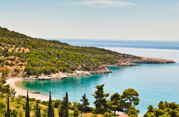 Scenic view of a secluded beach in Spetses, Greece. This tranquil bay features a narrow strip of sandy beach flanked by lush greenery and Mediterranean pines, leading into clear turquoise waters. The bay is surrounded by rocky outcrops and gentle hills, creating a private and serene atmosphere ideal for relaxation and swimming.