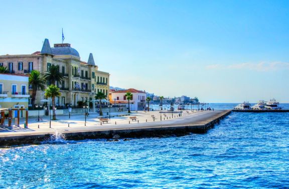 Bright and lively waterfront promenade in Spetses, Greece, featuring historic architecture with elegant facades and pointy rooftops. The wide pedestrian walkway along the sea is dotted with benches, leading to a dock lined with moored yachts. The scene captures a vibrant blue sea under a clear sky, emphasizing the charming, inviting atmosphere of this picturesque island destination.