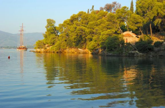 Old classic sailing yacht anchored at Love Bay on Poros Island in Greece, with green shore full of pine trees and a small old stone church