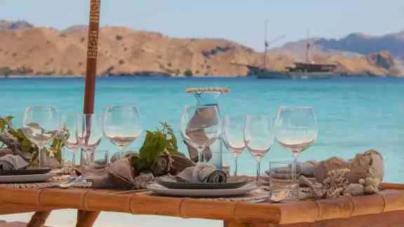 Seaside dining table set with wine glasses and a yacht anchored in a bay in Greece.