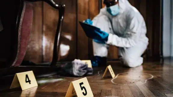 Investigator in protective gear examining a room with evidence markers on the floor.