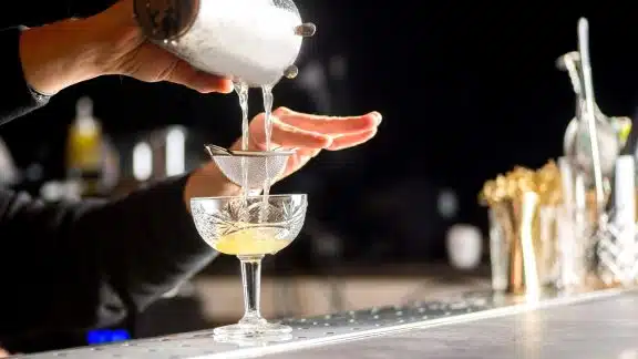 Cocktail being poured into a coupe glass at a bar.