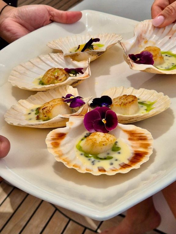 M/Y MILOS AT SEA serves Scallops in Shell Perfectly seared scallops in their natural shells, finished with a light citrus butter and edible flowers—almost too pretty to eat.
