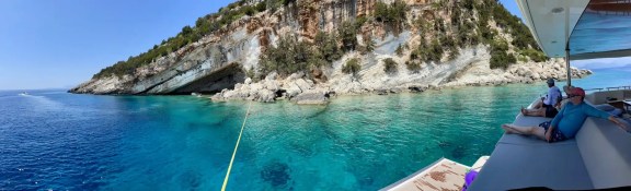Guests relaxing on the aft deck of a yacht beside a Greek island cove.