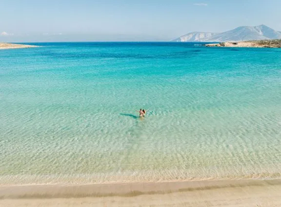 Aerial view of swimmers in the shallow turquoise water of a sandy Koufonisia beach, with Keros island in the background